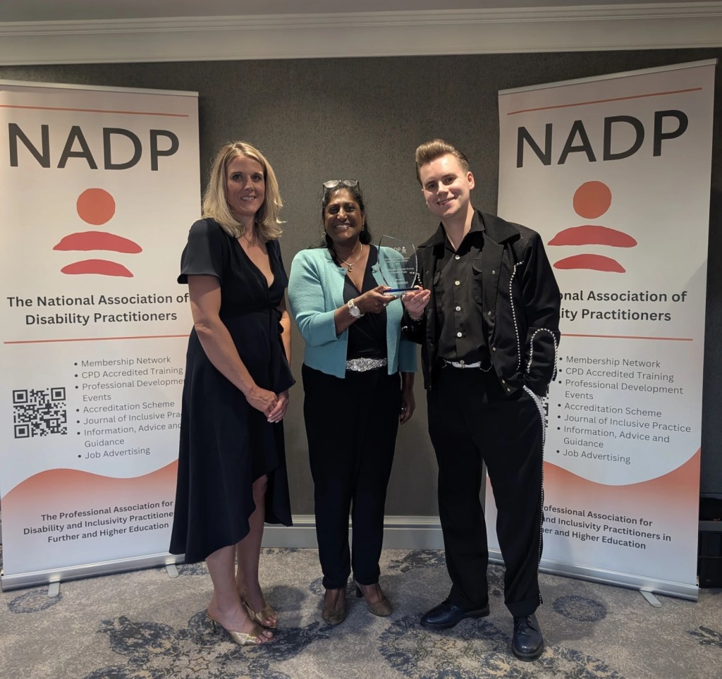 Three people posing with an award at an NADP conference, standing between two organisational banners.