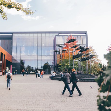 Students walking across a modern university campus plaza with glass buildings and sculptural installation.