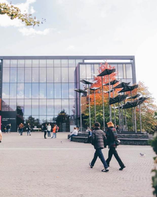 Students walking across a modern university campus plaza with glass buildings and sculptural installation.