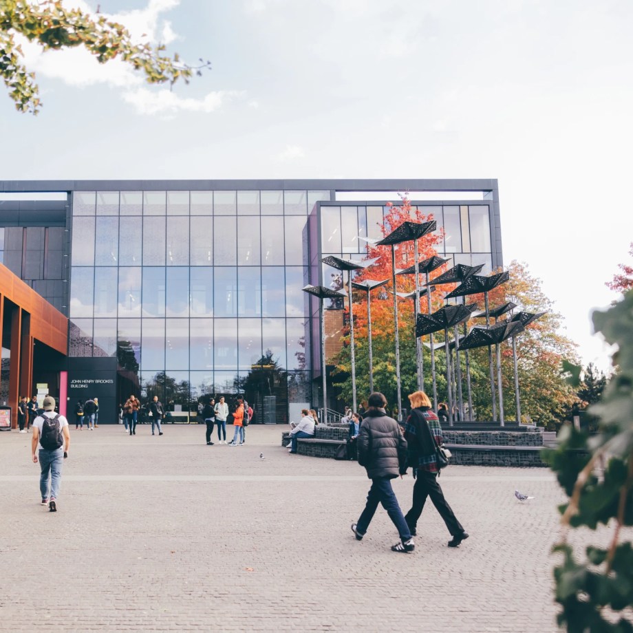 Students walking across a modern university campus plaza with glass buildings and sculptural installation.