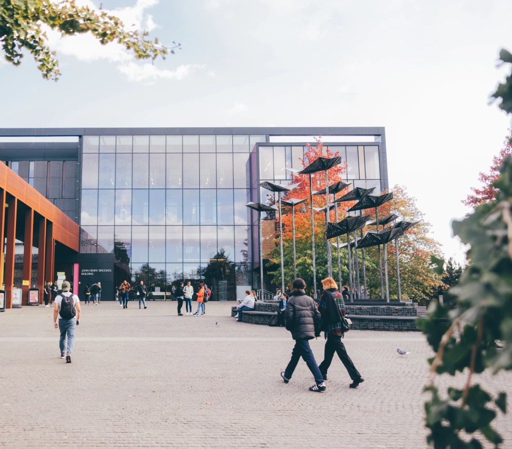 Students walking across a modern university campus plaza with glass buildings and sculptural installation.