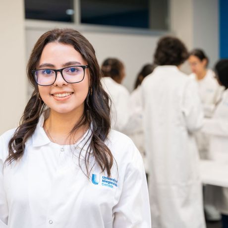 Young woman with glasses and long dark hair smiling in a white lab coat, with other lab-coated students working in the background.