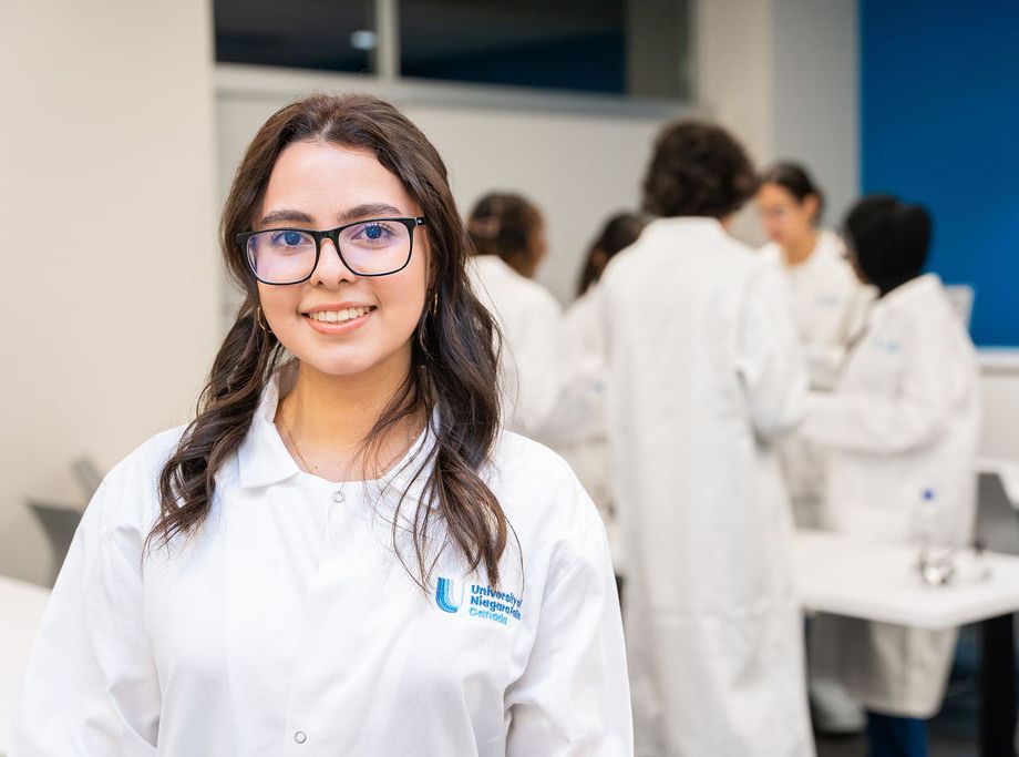 Young woman with glasses and long dark hair smiling in a white lab coat, with other lab-coated students working in the background.