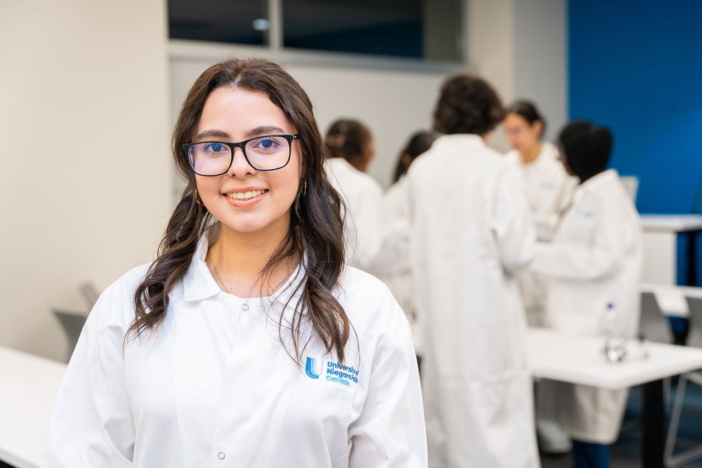 Young woman with glasses and long dark hair smiling in a white lab coat, with other lab-coated students working in the background.