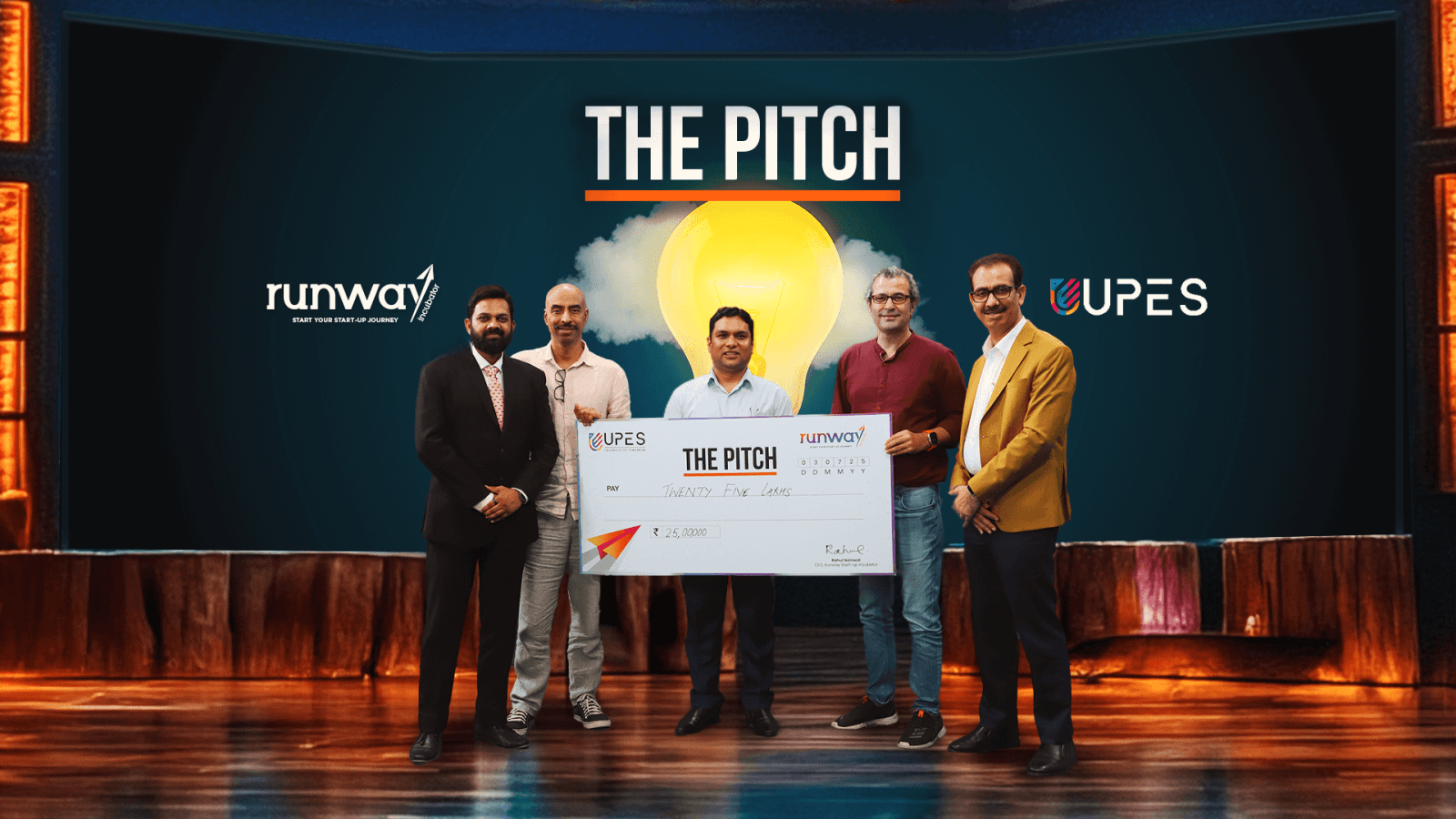 Five men holding large ceremonial check at 'The Pitch' startup competition event with runway and UPES branding.