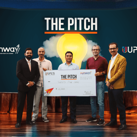 Five men holding large ceremonial check at 'The Pitch' startup competition event with runway and UPES branding.