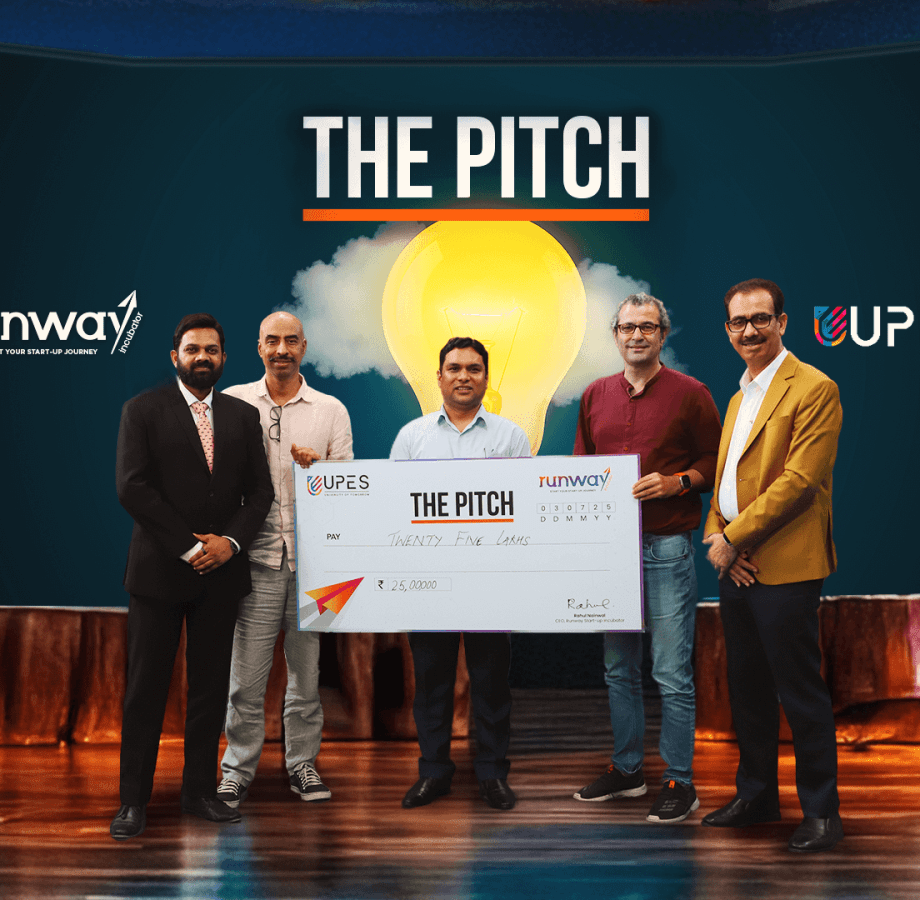 Five men holding large ceremonial check at 'The Pitch' startup competition event with runway and UPES branding.