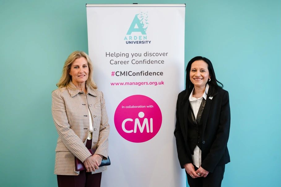 The Duchess of Edinburgh standing with an Arden University staff member in front of an Arden University and CMI banner against a turquoise wall.