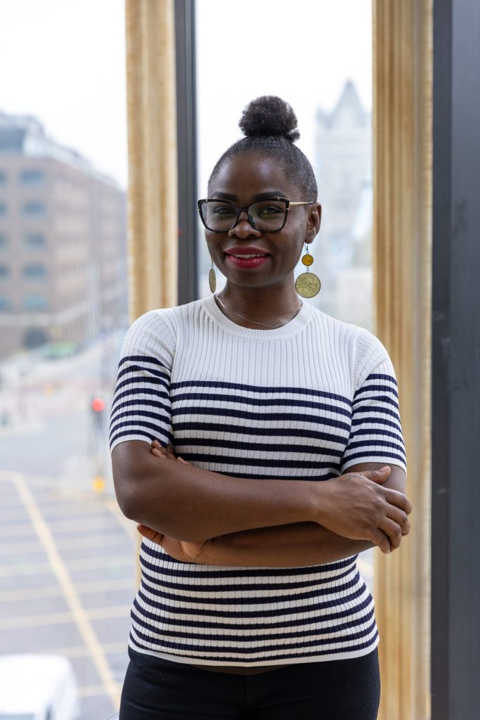 Smiling woman in glasses and striped top standing by a window with city view