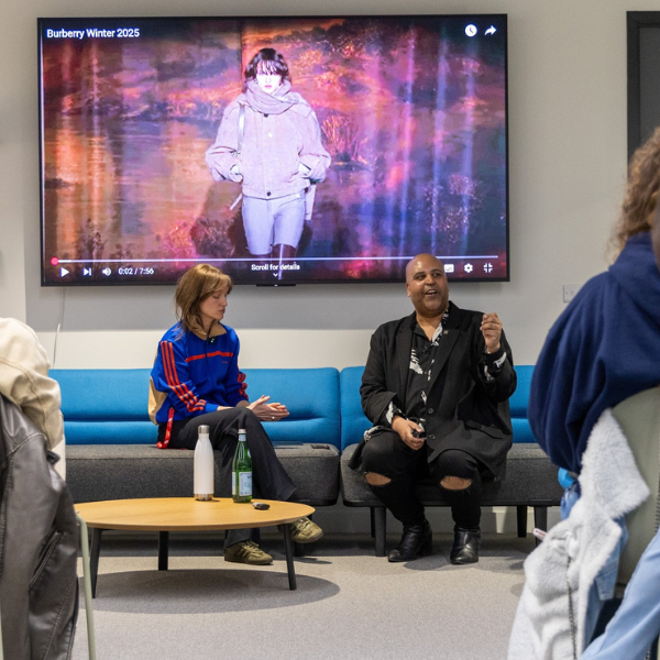 Two people seated on a blue couch in a modern room, engaged in conversation. Behind them, a large screen displays a video titled 'Burberry Winter 2025,' showing a model walking on a runway in winter fashion.