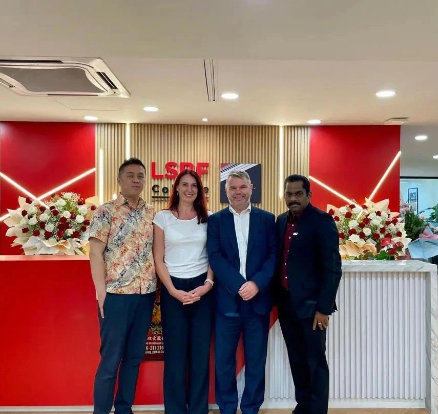 Three men and one woman standing in front of a reception desk with floral arrangements, against a red and white backdrop featuring the LSBF College logo.