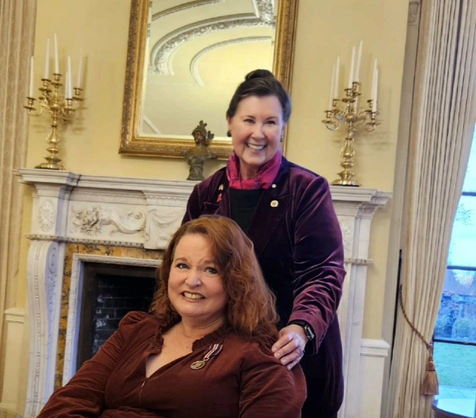 Two women in dark formal attire, one standing and one sitting and wearing a Coronation Medal, posed in a formal room with an ornate fireplace and tall windows.
