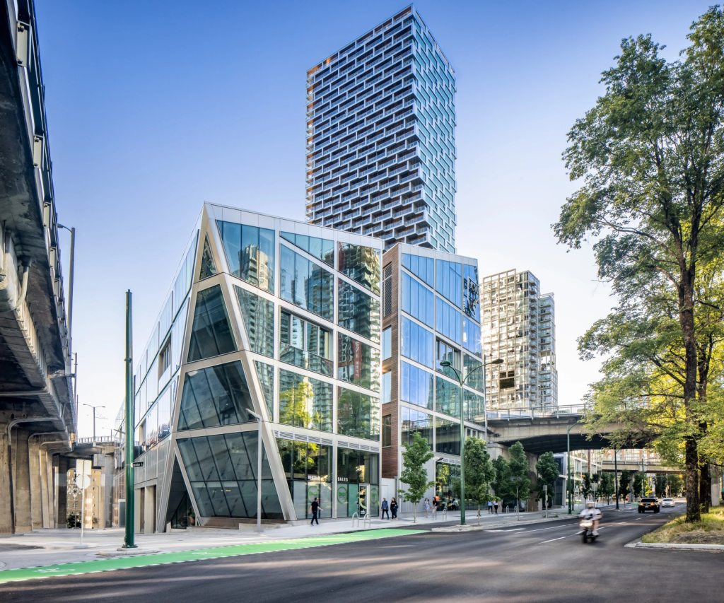Modern glass building of the UCW campus in Vancouver, with high-rise towers and trees nearby.