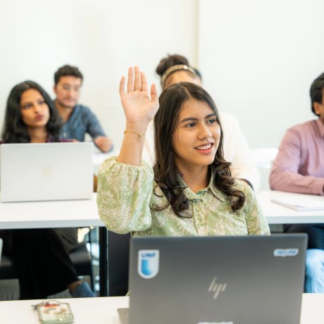 Student raising hand in classroom with laptops and other students in background.