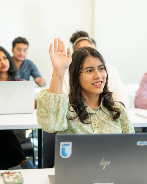 Student raising hand in classroom with laptops and other students in background.