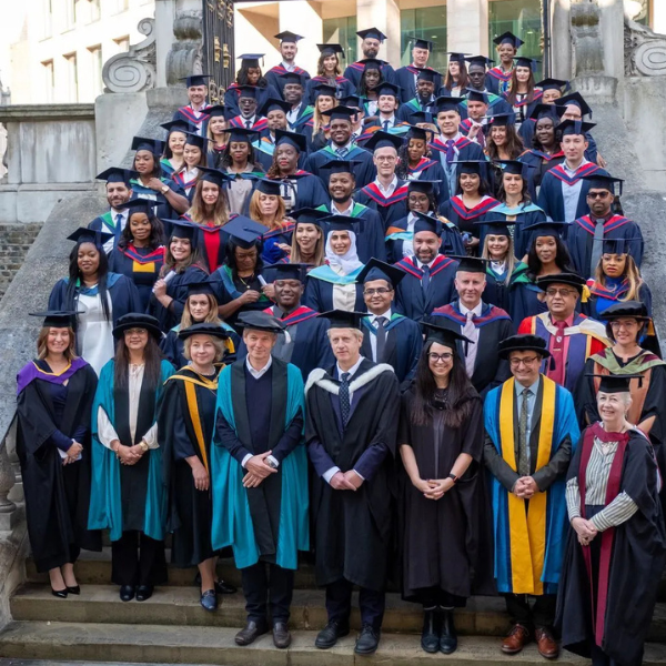 Large group of graduates in academic gowns and caps posing on outdoor steps with faculty members in formal attire.