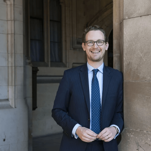 Person in a pinstripe suit smiling in front of a historic stone building with arched windows.