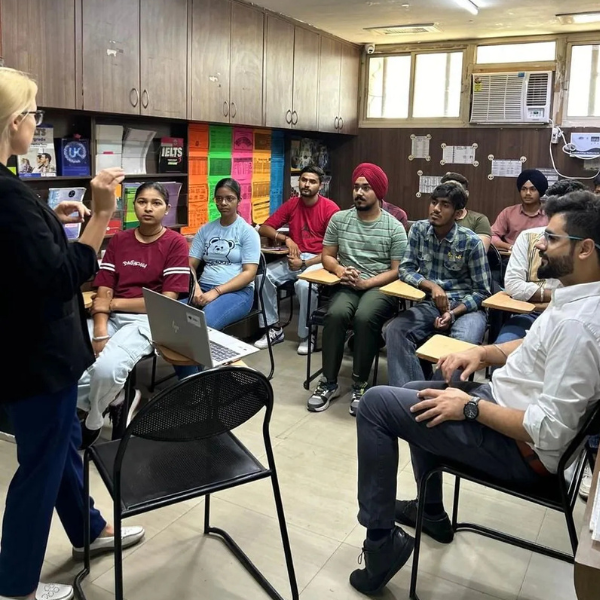 Instructor teaching a group of students in a classroom with desks, posters, and a laptop.