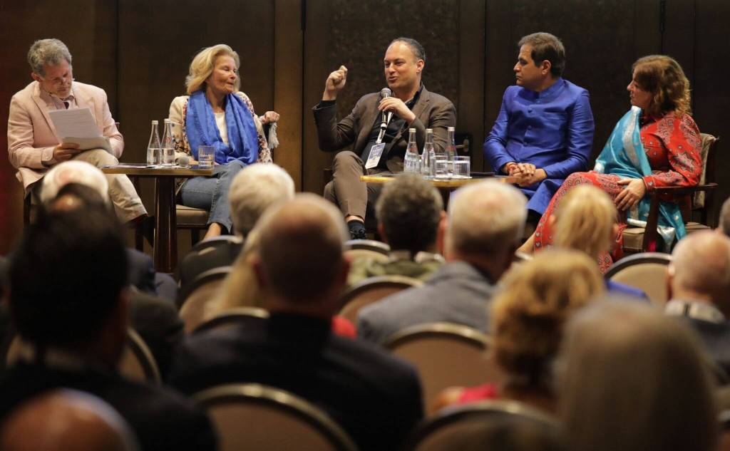 Five speakers sit onstage during a panel discussion, addressing an audience in a conference room.