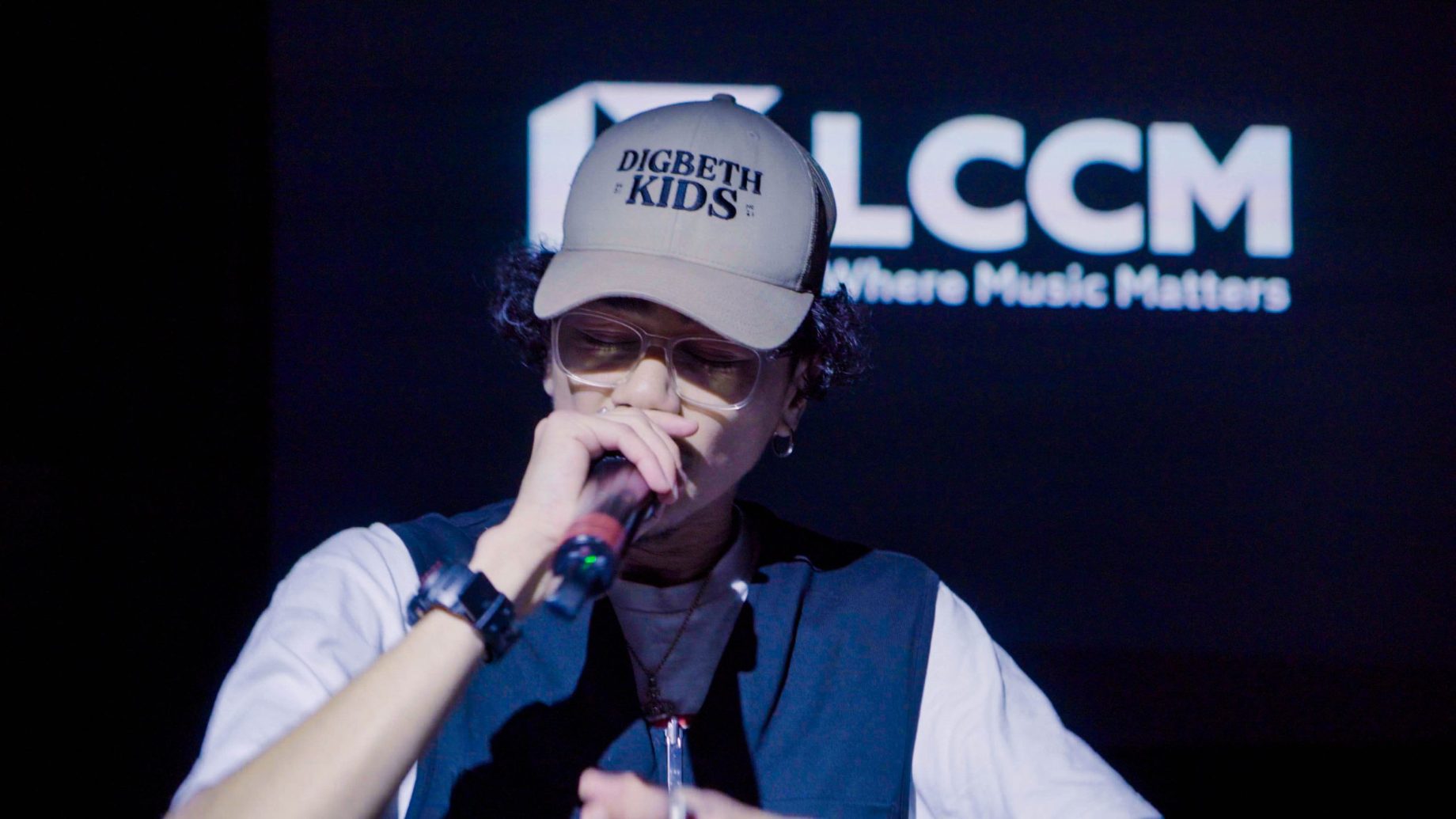Person in a 'DIGBETH KIDS' cap holding a microphone in front of an LCCM backdrop