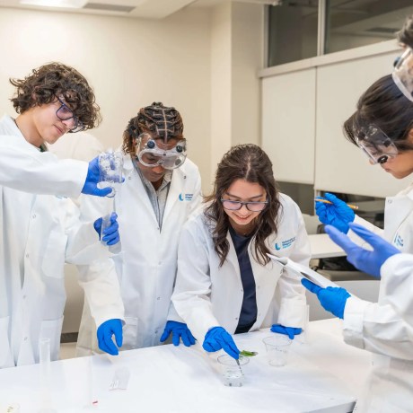 Four students in lab coats and safety goggles conducting a science experiment together in a laboratory.