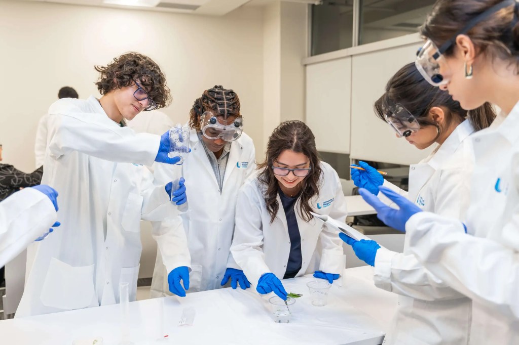 Four students in lab coats and safety goggles conducting a science experiment together in a laboratory.