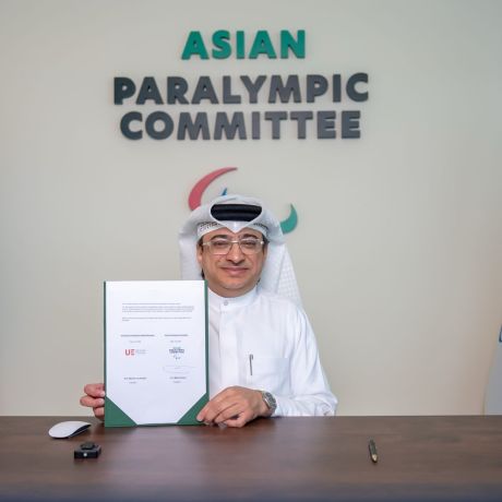 A representative of the Asian Paralympic Committee sits at a desk holding a signed agreement document, with the University of Europe for Applied Science in Dubai and Asian Paralympic Committee flags displayed on either side and the organisation’s logo in the background.