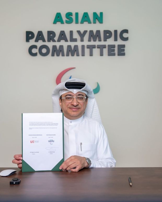 A representative of the Asian Paralympic Committee sits at a desk holding a signed agreement document, with the University of Europe for Applied Science in Dubai and Asian Paralympic Committee flags displayed on either side and the organisation’s logo in the background.
