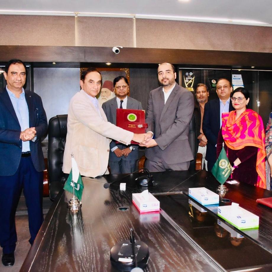 Group of university and institutional representatives gathered around a conference table during a memorandum of understanding signing ceremony in Pakistan, with two men exchanging a commemorative plaque.